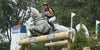 Side front view of a grey horse and its rider going over a jump