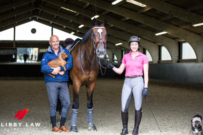 Man and woman stood either side of a horse and smiling. The man is holding a sausage dog.
